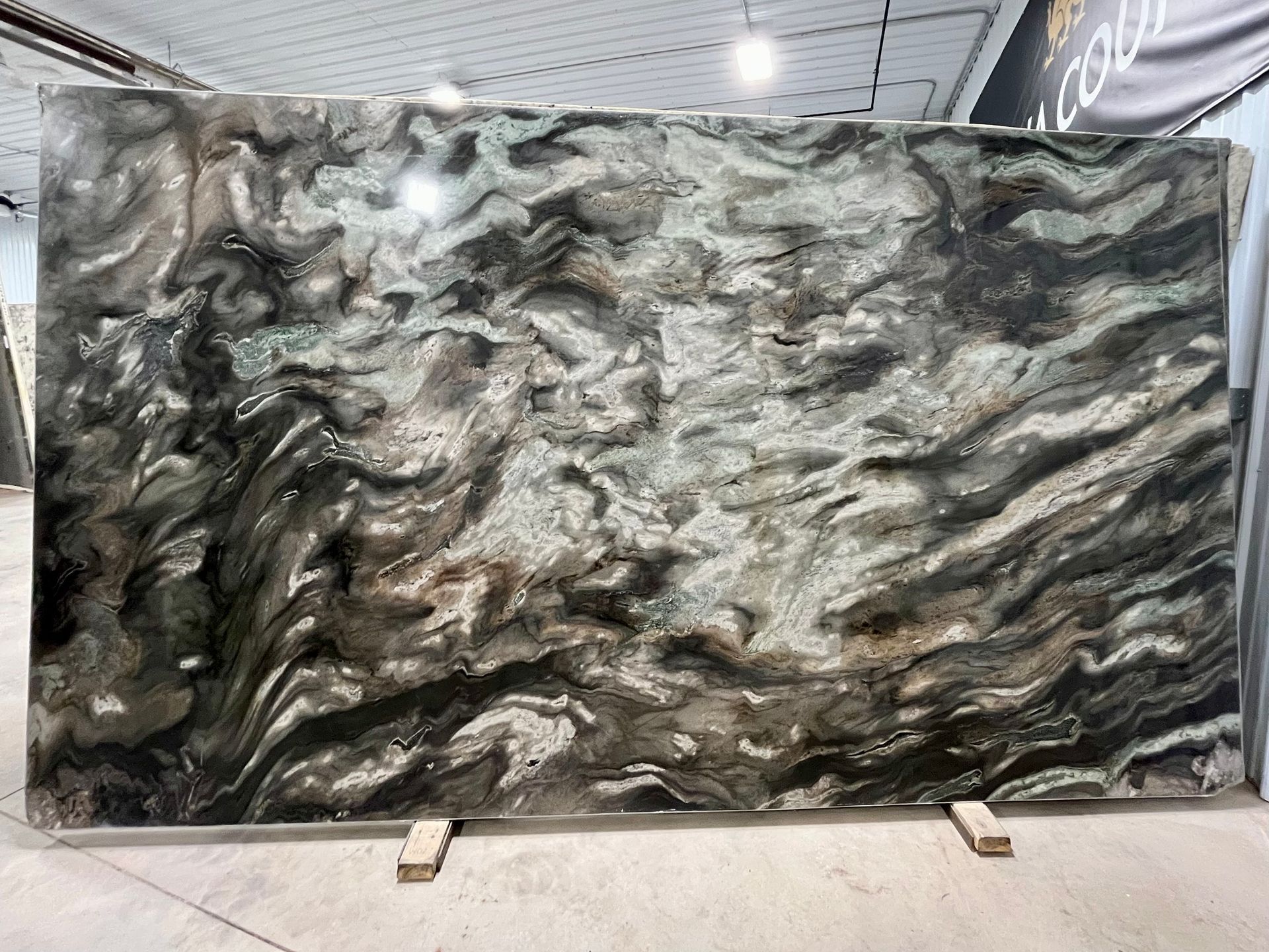 Large slab of polished stone with swirling patterns of black, grey, and white, displayed in a warehouse.