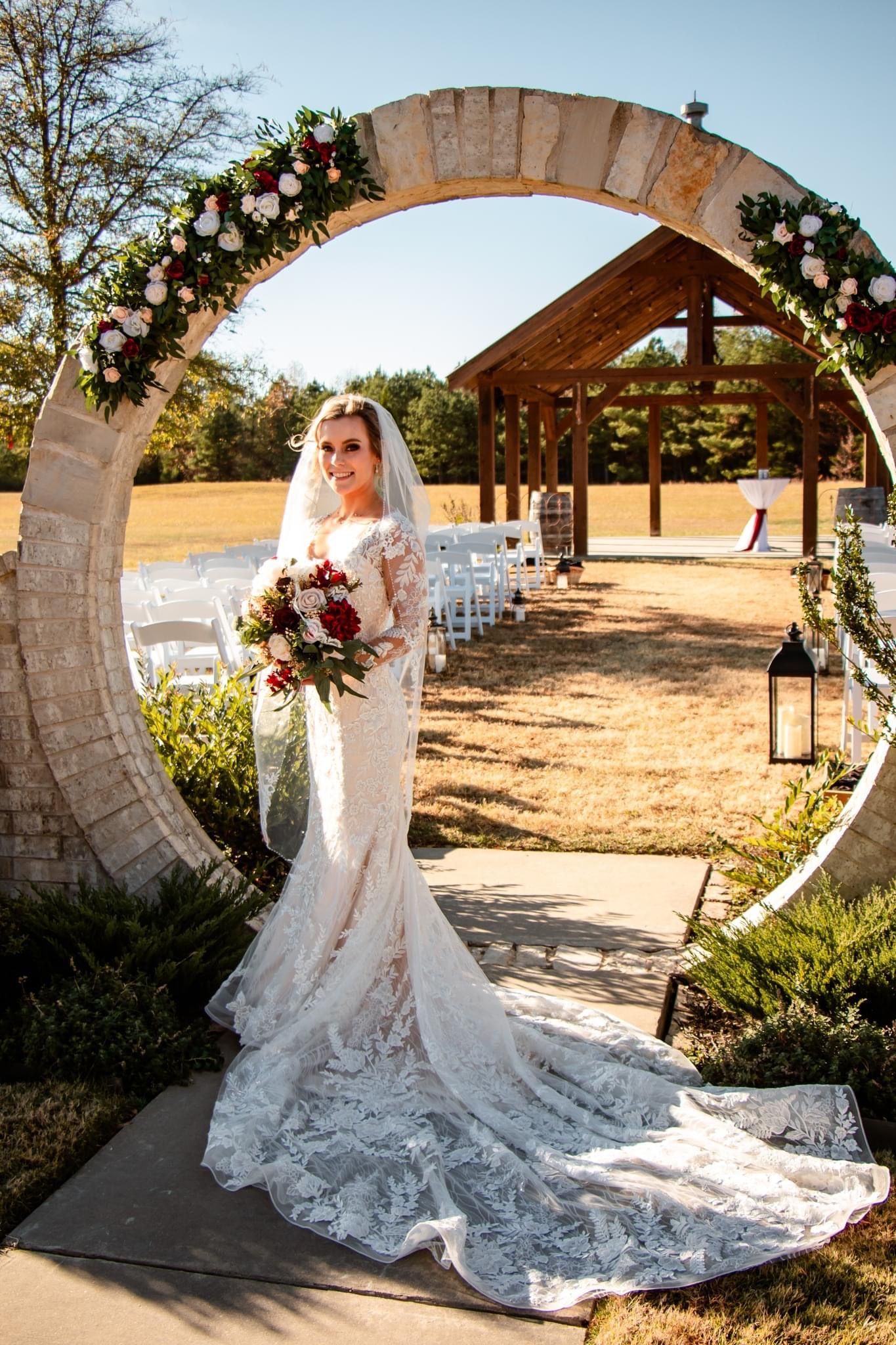 A bride in a wedding dress is standing in front of a stone arch holding a bouquet of flowers.