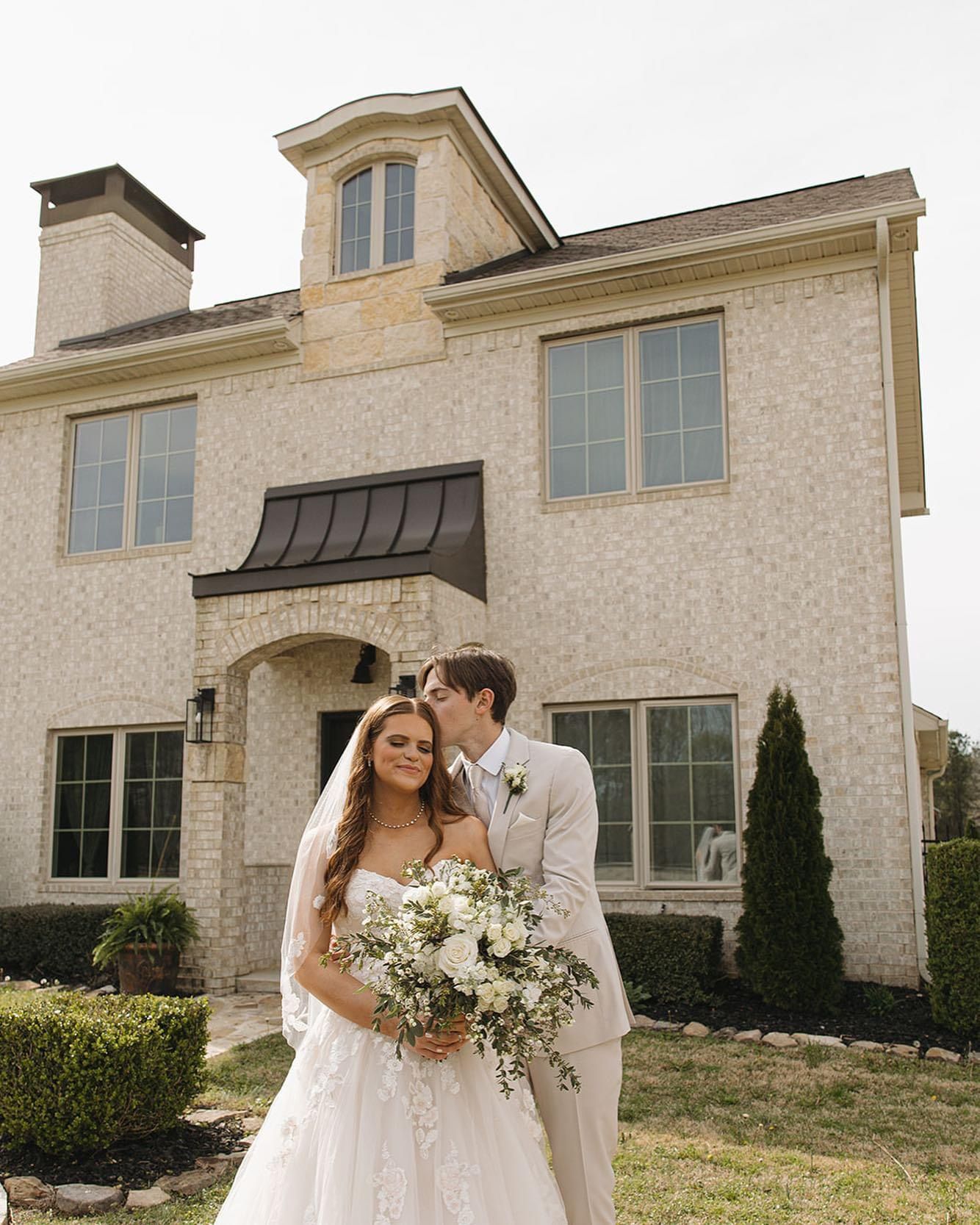 A bride and groom are kissing in front of a large house.