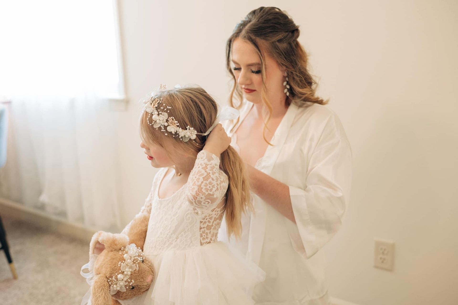 A woman is brushing a flower girl 's hair while holding a teddy bear.