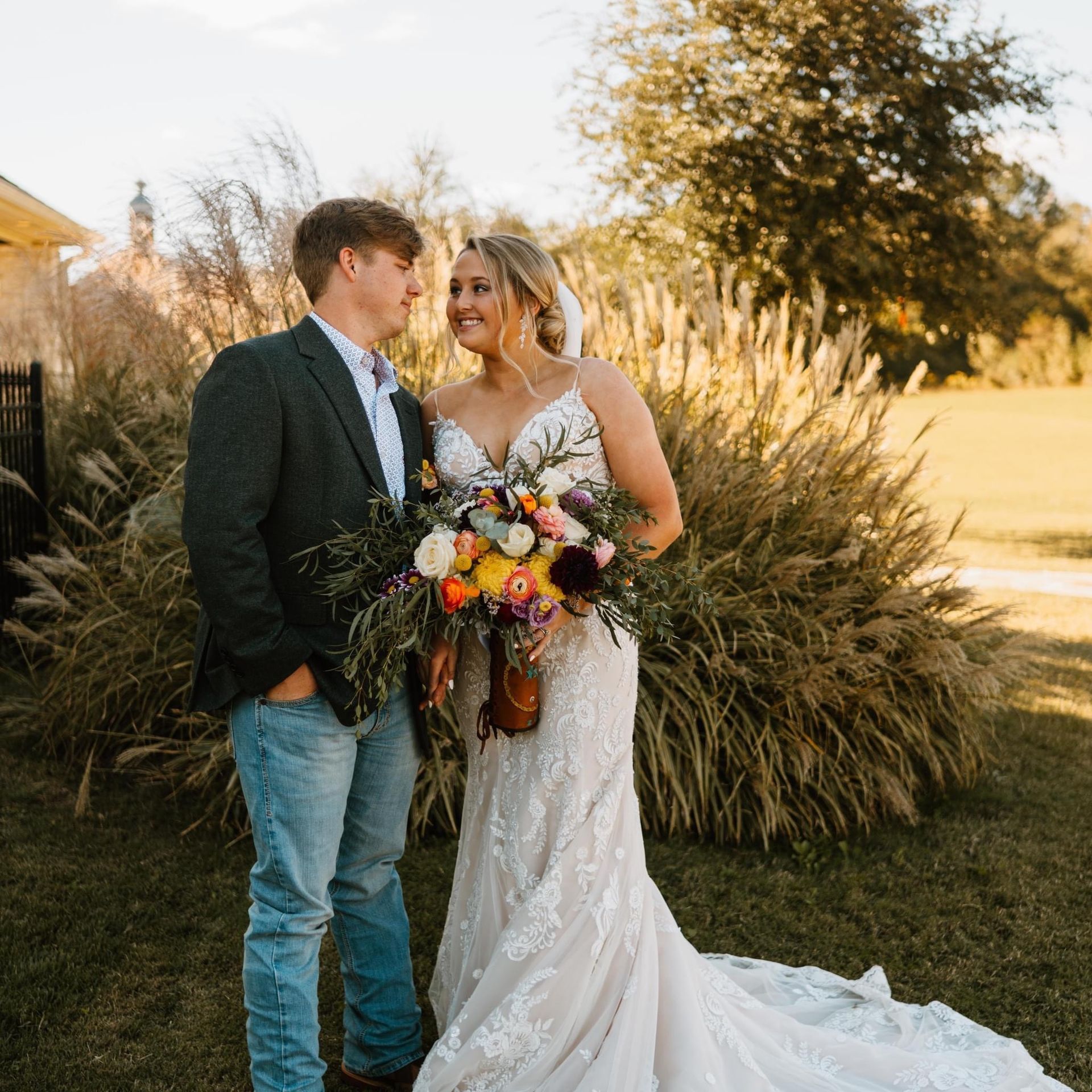 A bride and groom are standing next to each other in a field holding a bouquet of flowers.