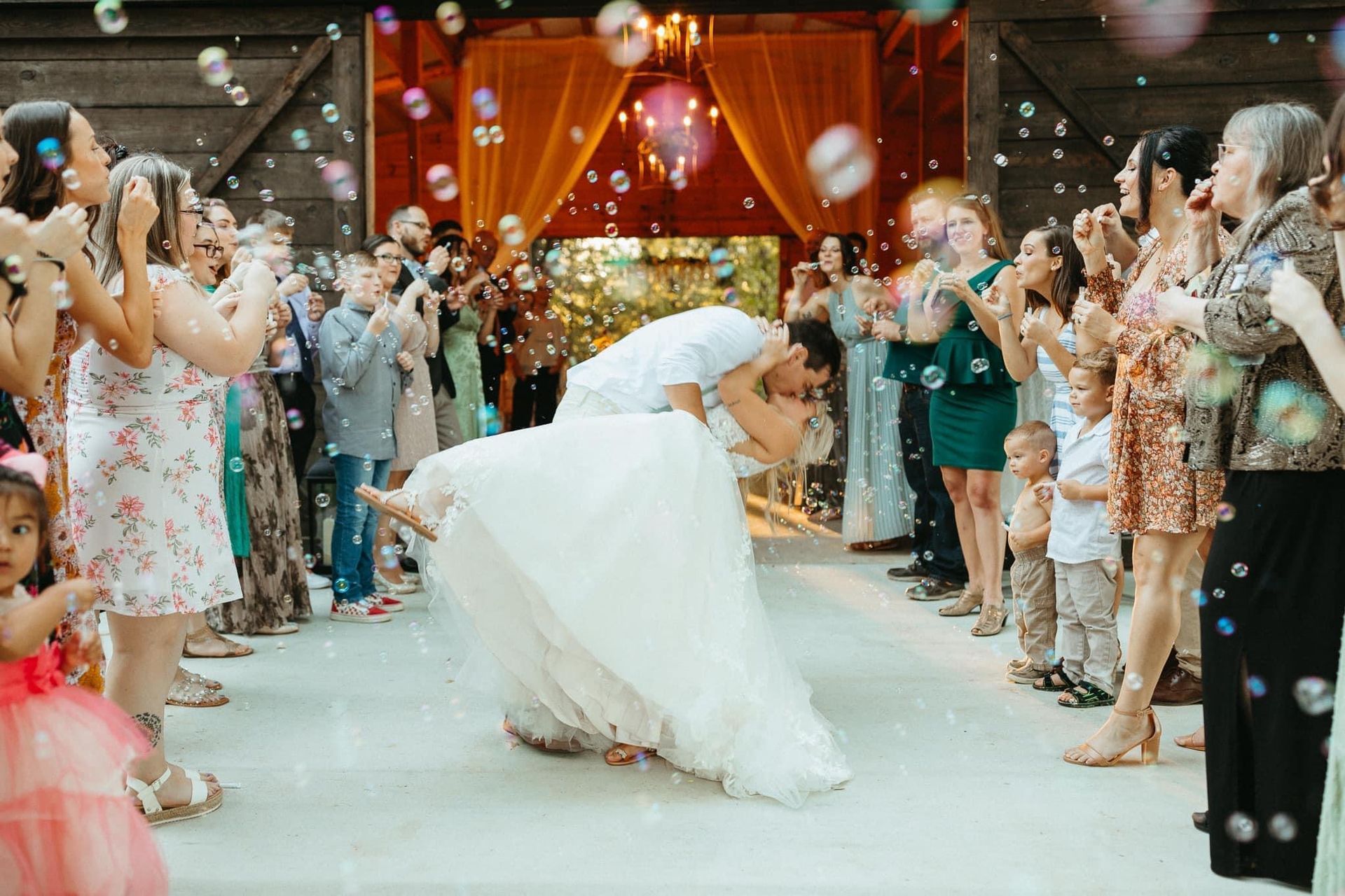 A bride and groom are kissing while their wedding guests blow soap bubbles.