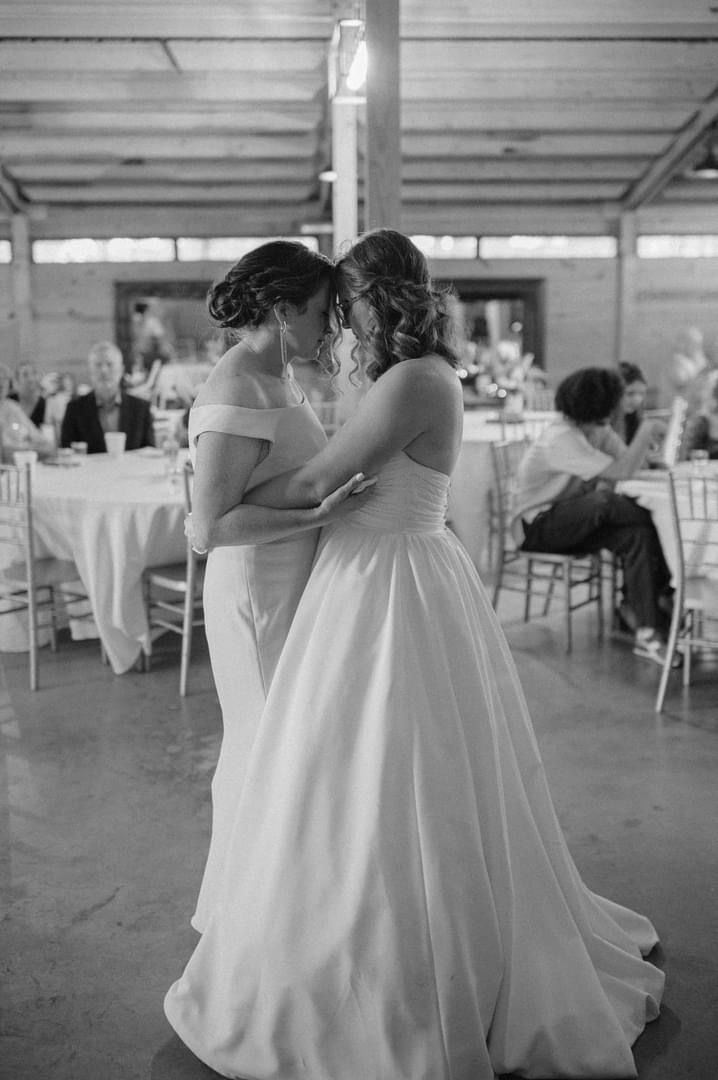 Two women in wedding dresses are dancing together in a black and white photo.