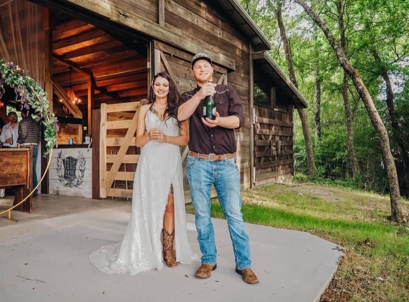 A bride and groom are standing in front of a barn holding a bottle of champagne.
