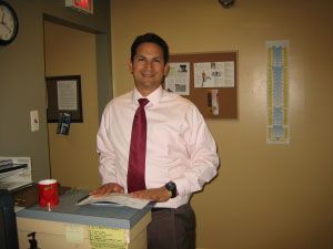 Man in pink shirt and tie at a desk, smiling. Office setting, documents on the counter.