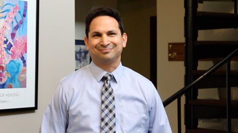 Man in blue shirt and tie smiles, standing by stairs and artwork.