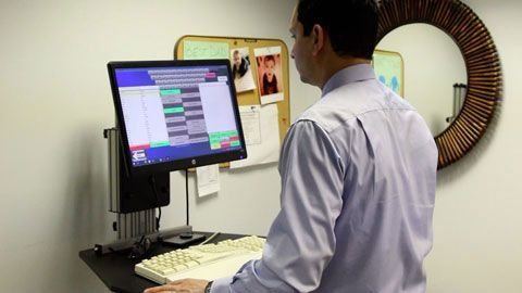 Man at standing desk, viewing computer screen with data display. Office setting.