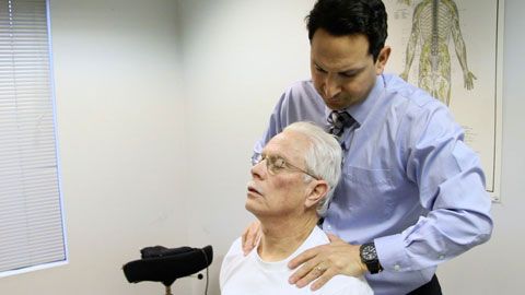 Chiropractor examining a patient's neck, applying pressure. White room, anatomical chart on the wall.