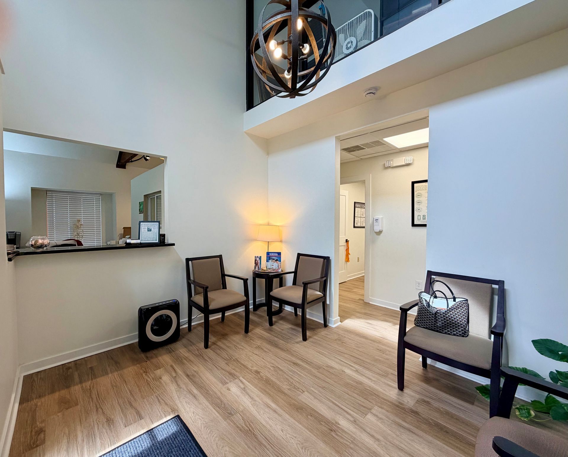 A bright waiting room featuring wood-style flooring, three armchairs, a central side table with a lamp, and a tall ceiling.