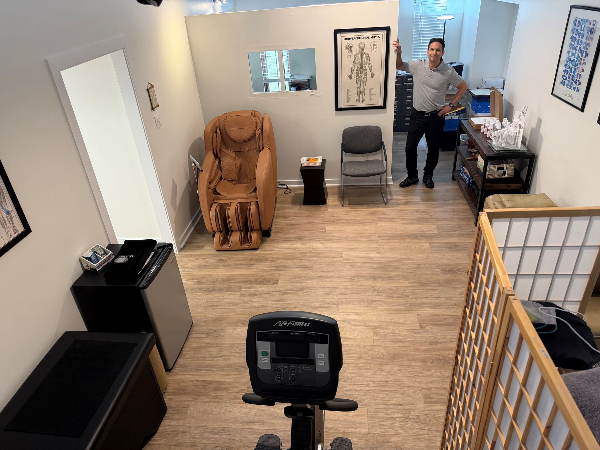 A man stands in a room with a massage chair, an exercise bike, a chair, and a framed anatomical chart on the wall.