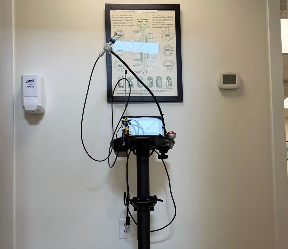 Medical device mounted on a wheeled stand in a clinic room, with cords and a small display screen.