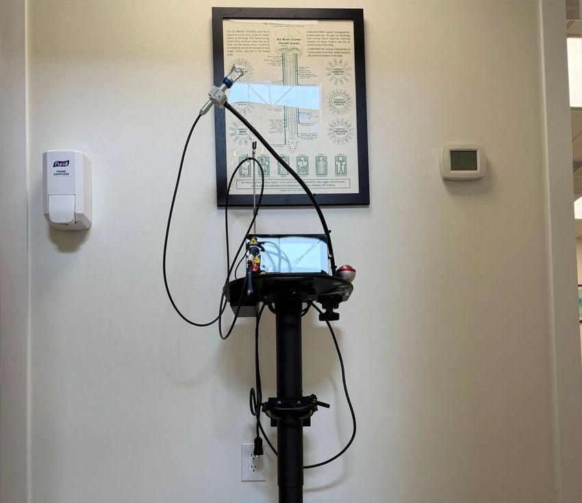Medical device mounted on a wheeled stand in a clinic room, with cords and a small display screen.