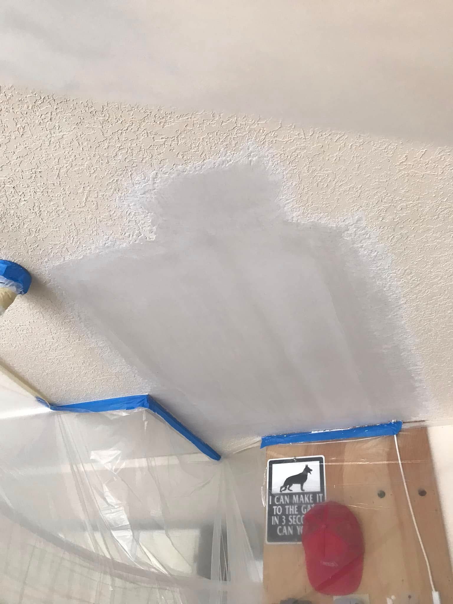 Ceiling being painted, blue tape marks a border. Plastic sheeting protects surrounding areas.