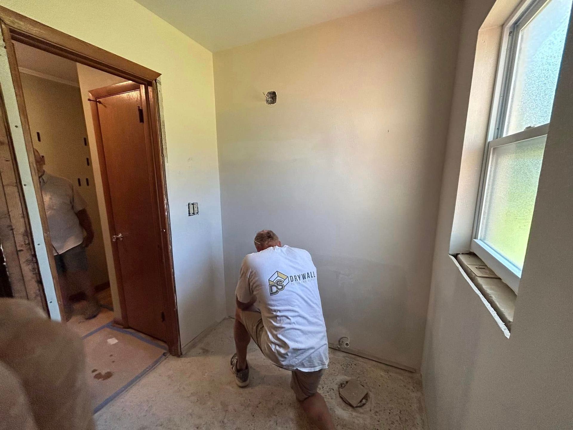 Man kneeling, working on a wall in a room with a window and doorway.