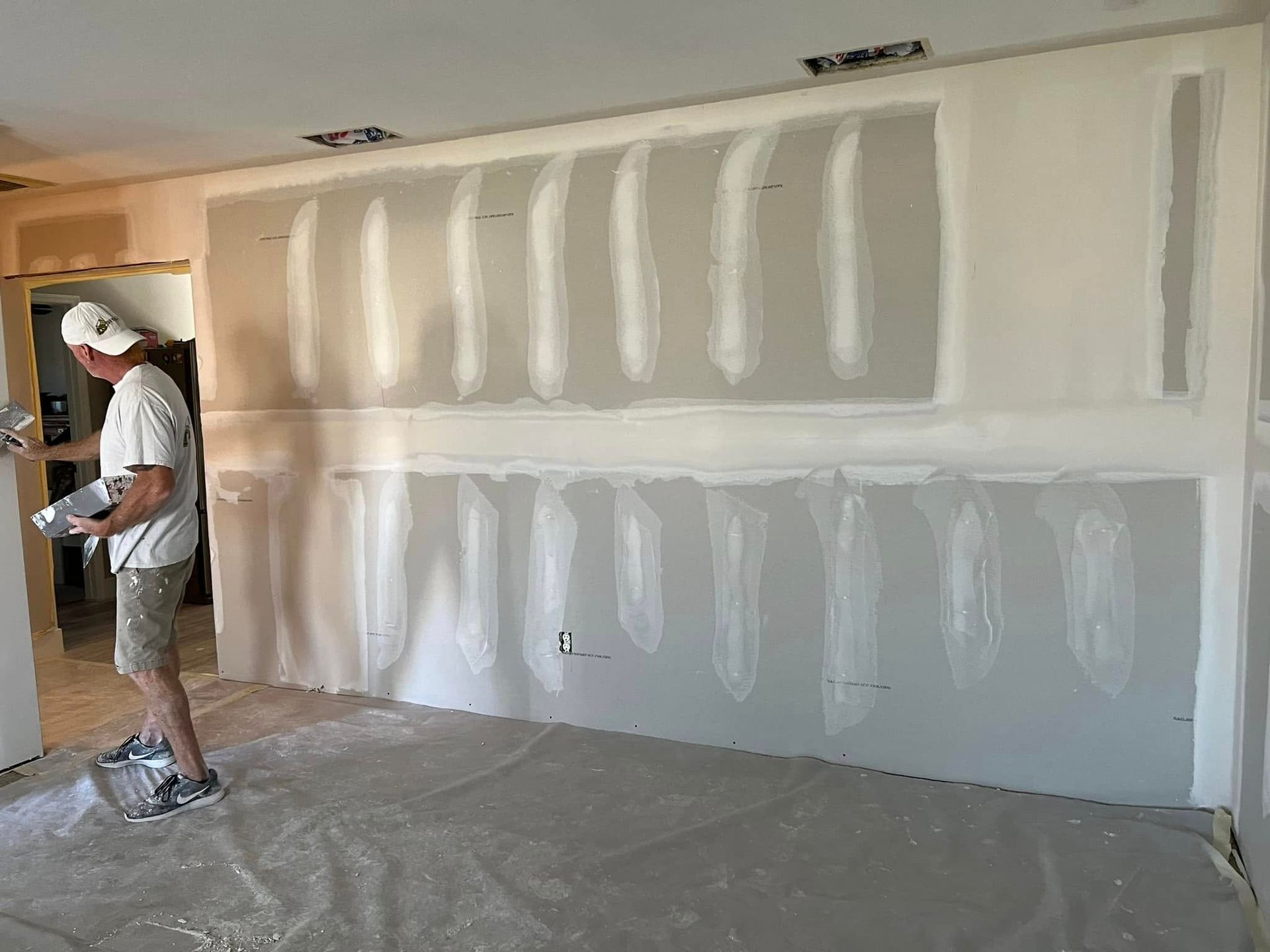Man applying drywall mud to a wall. Room under construction. White wall, gray floor covering.
