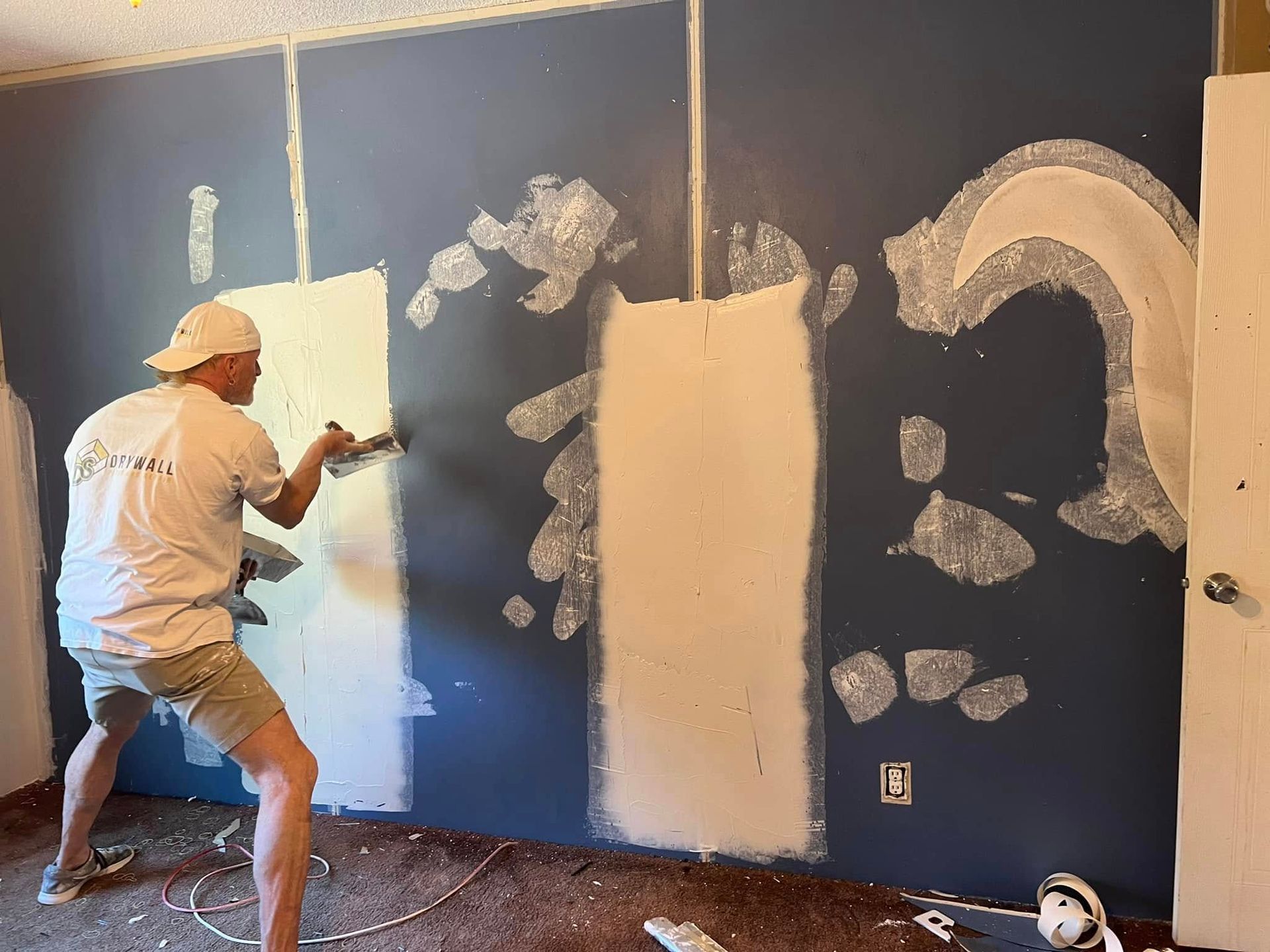 Person spraying white paint on a blue wall. Room with exposed drywall and electrical outlet.