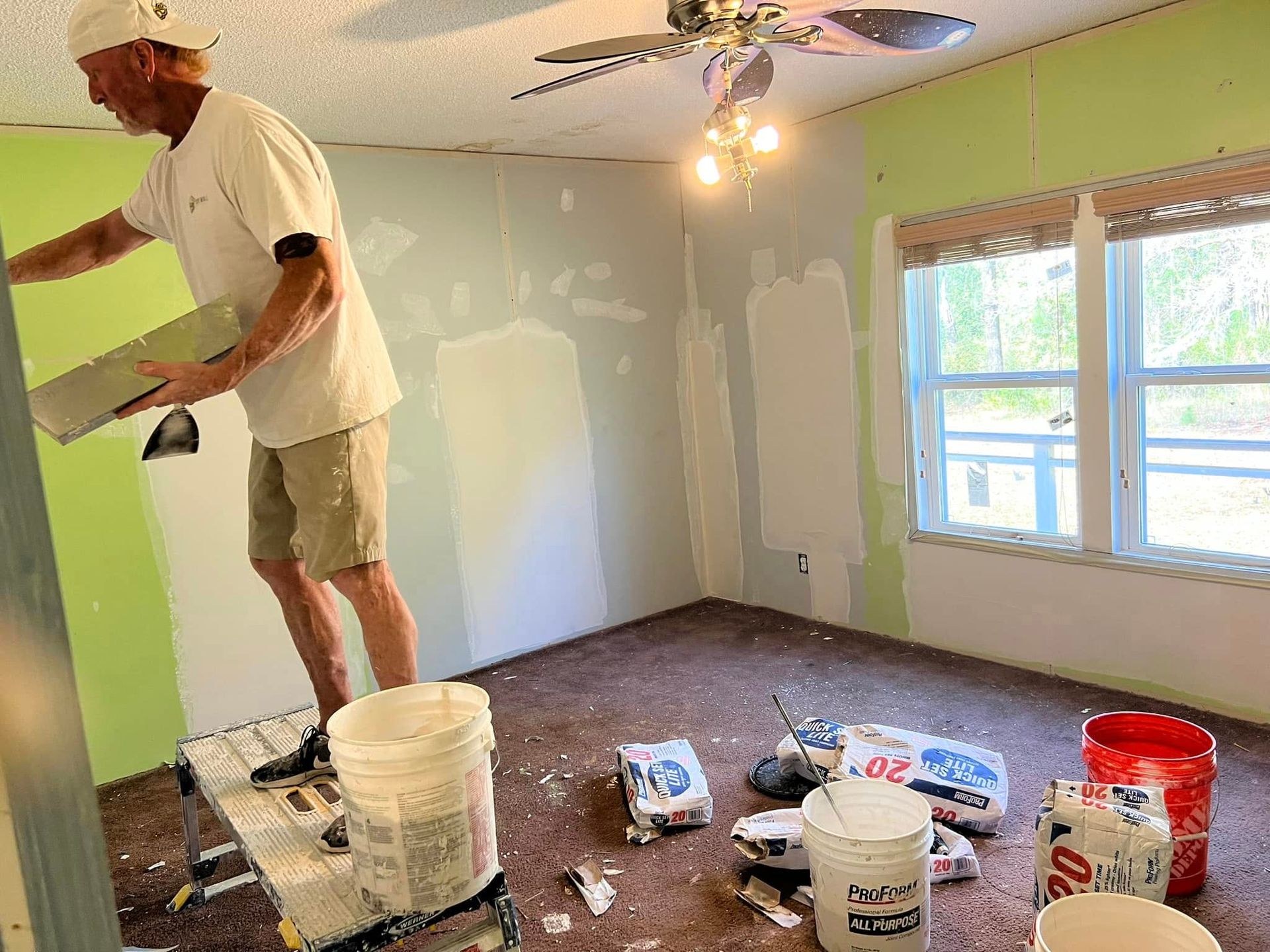 Man applying drywall mud in a room with green and white walls, buckets, and a window.