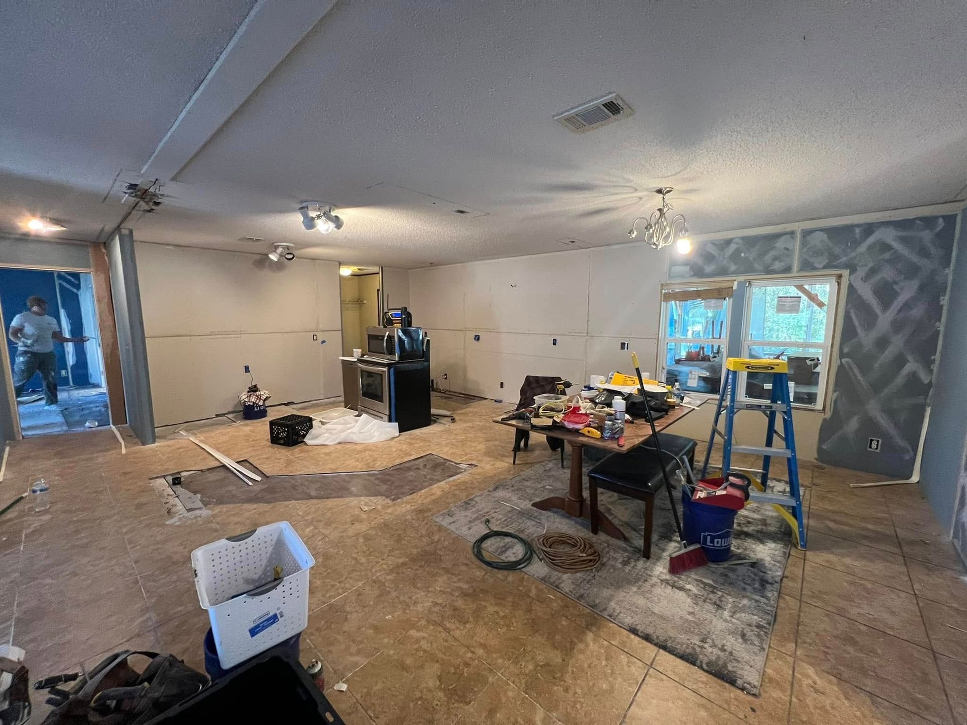 Interior view of a kitchen under renovation, drywall installed, tools and debris scattered about. Two people present.