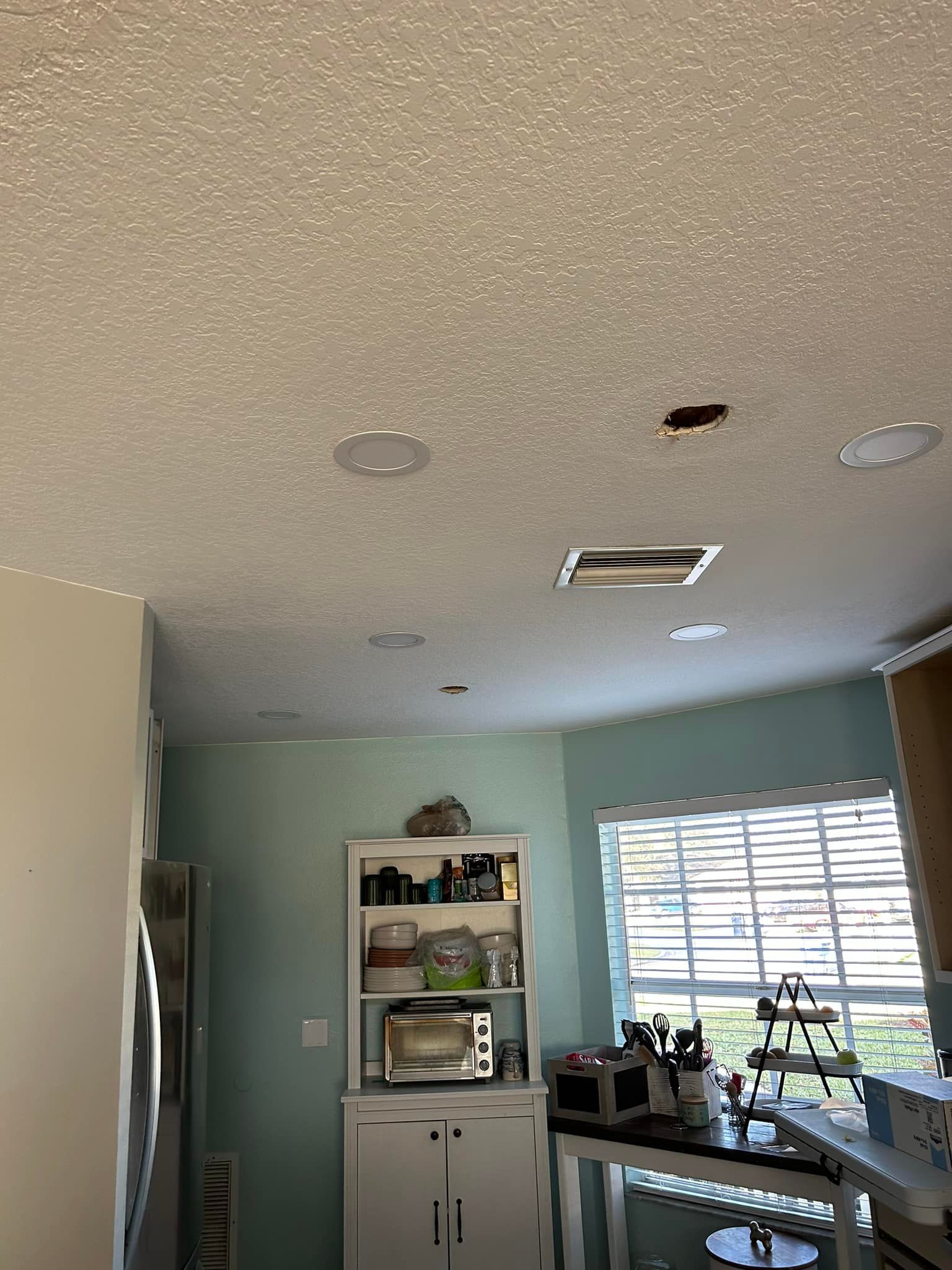 Kitchen ceiling with recessed lights, hole, and textured surface. Light blue walls and white cabinets.