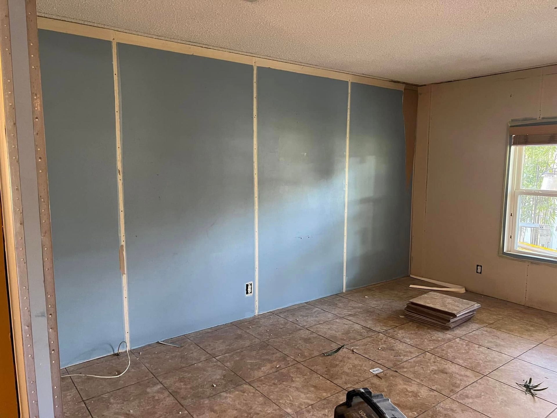 Room with unfinished blue-painted drywall on one wall, exposed wooden studs, and damaged flooring. Window visible.