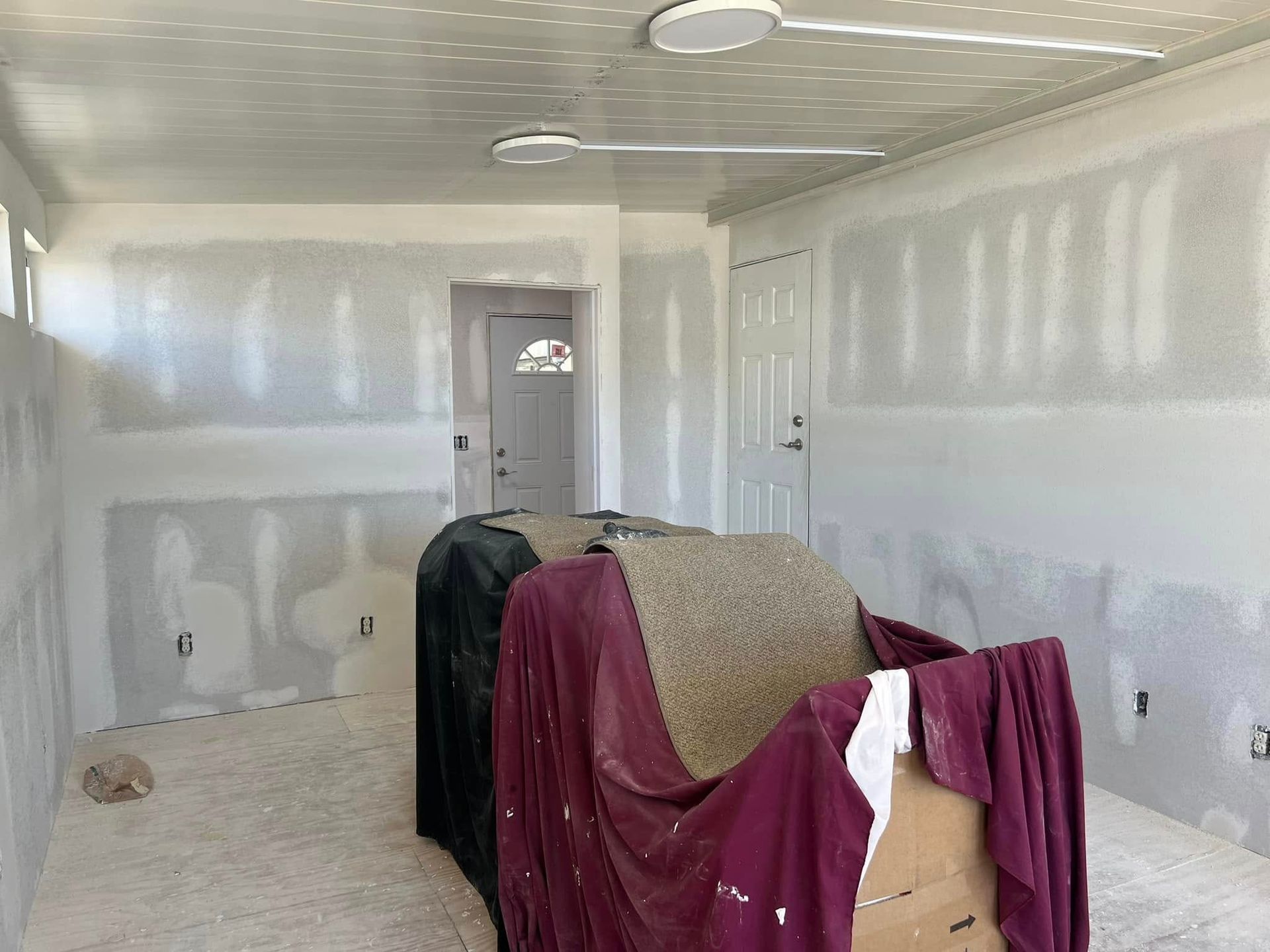 Room under renovation with drywall, covered furniture, and two ceiling lights.