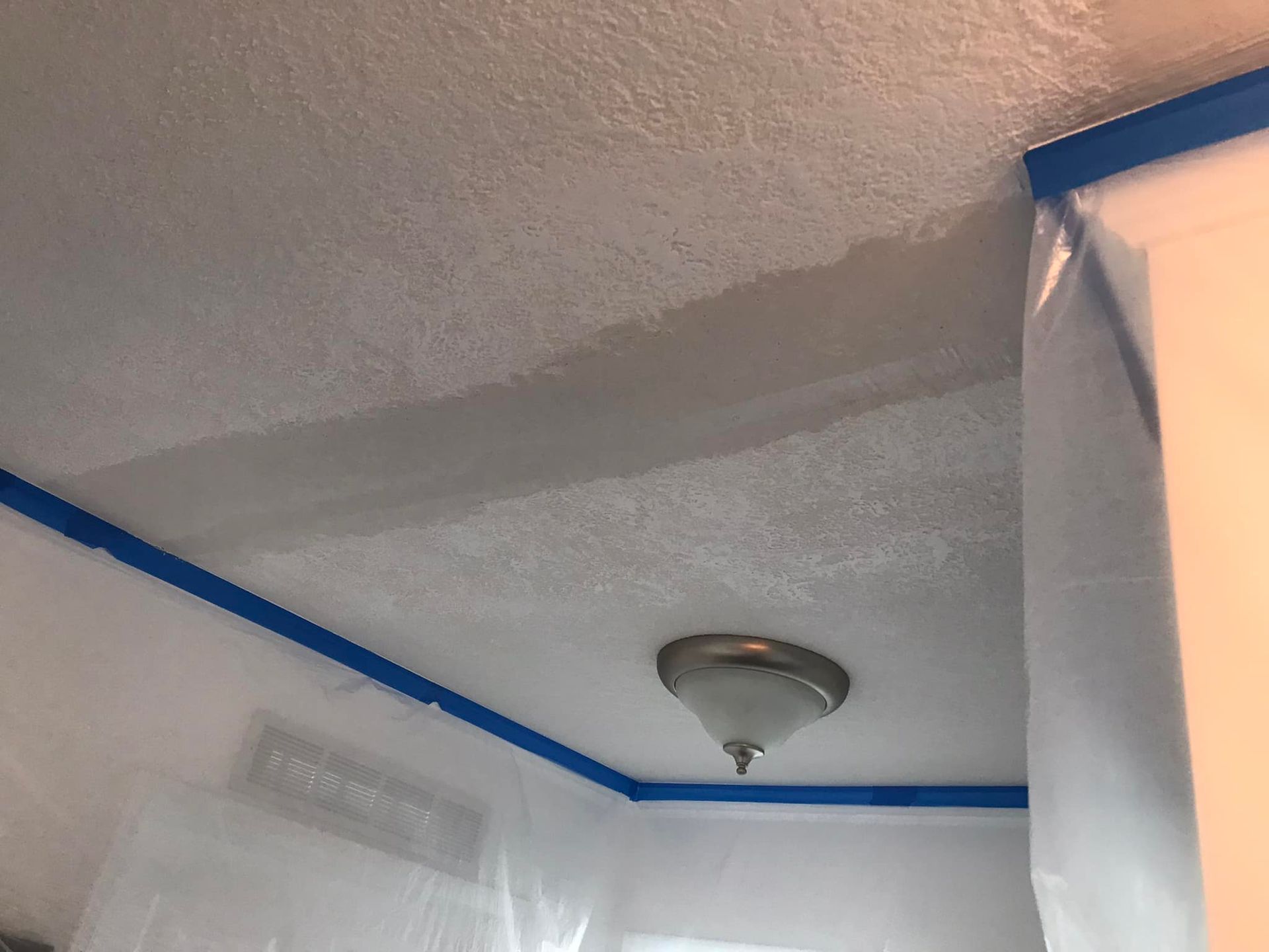 Ceiling being painted, blue painter's tape along edges. White textured ceiling with a light fixture.