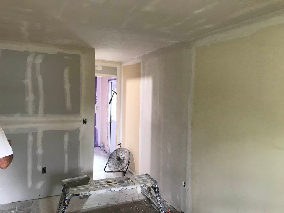 Room with drywall installation in progress; walls are primed, a ladder and fan are present.