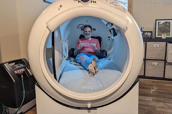 A man is sitting in an oxygen chamber on a bed.