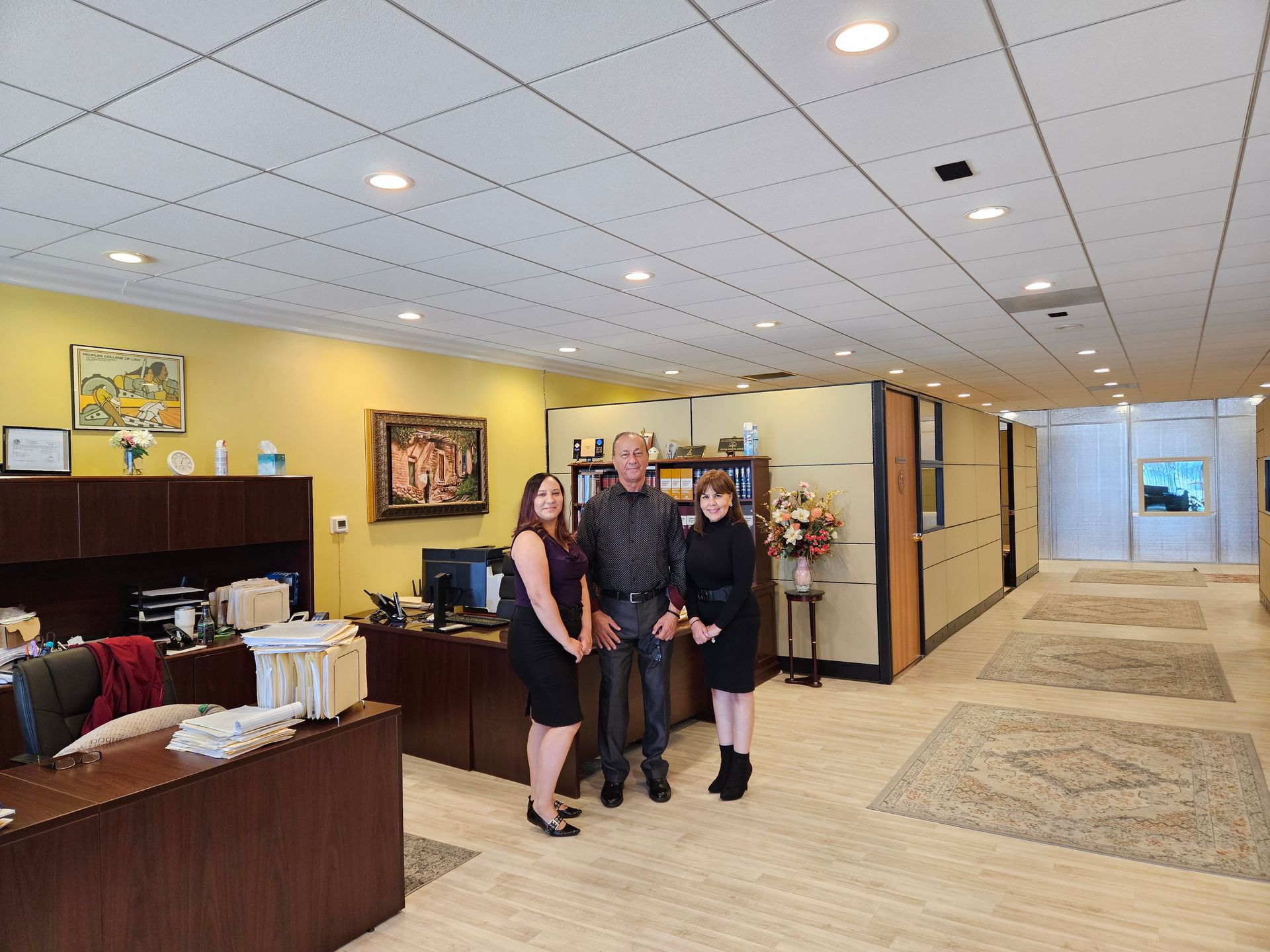 Three people standing in a hotel lobby near the front desk, with yellow walls and a tiled ceiling.