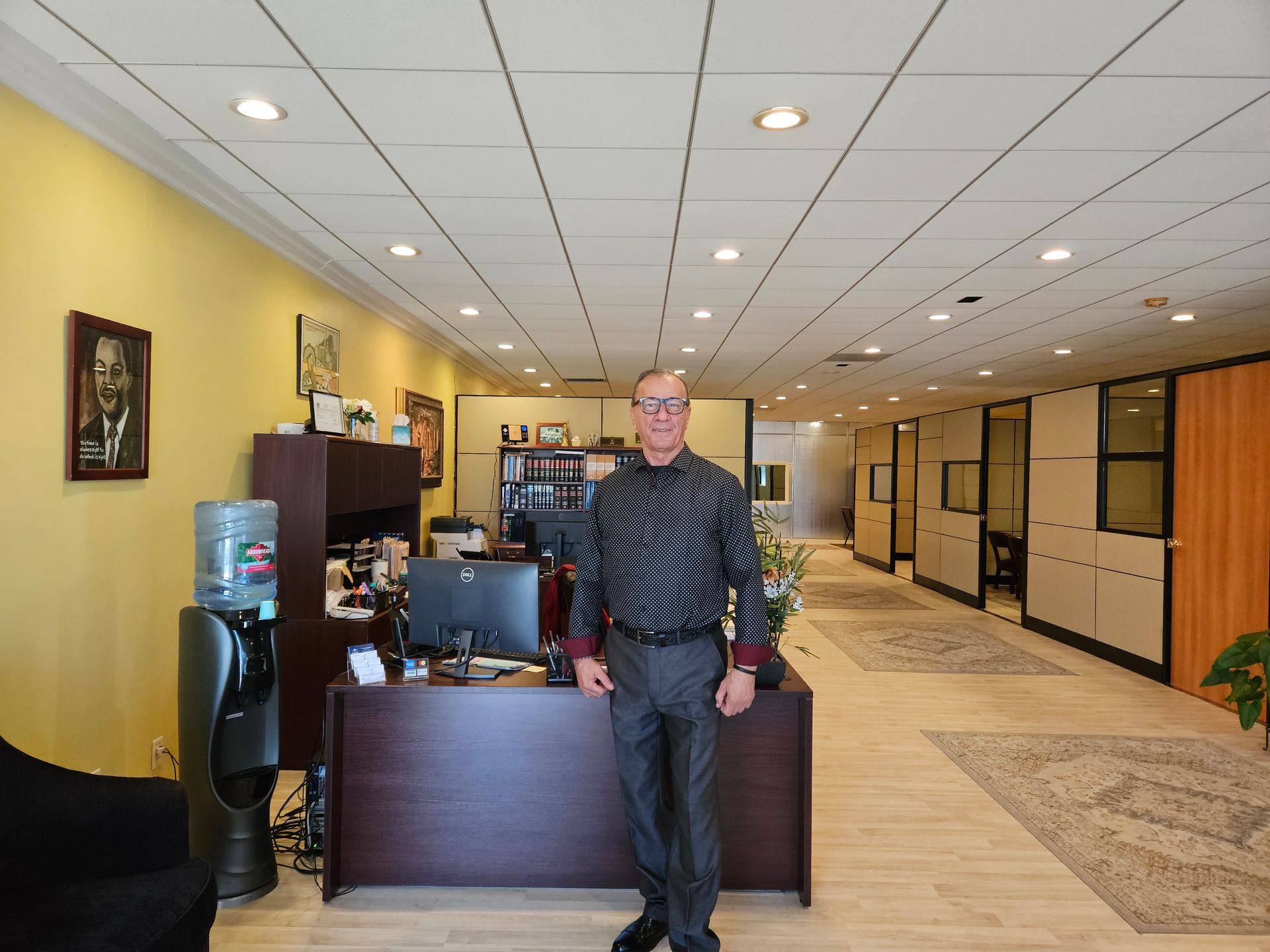 Man standing behind a front desk in a bright office lobby with carpeted hallway and framed art.