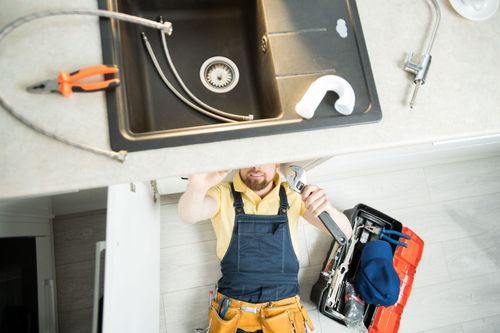 Worker in yellow shirt and overalls under a sink, holding tools in a utility room.
