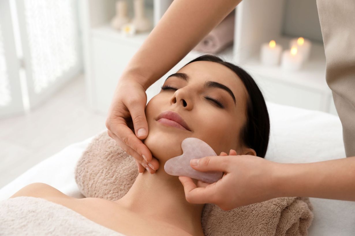 A practitioner performs a facial gua sha massage on a person using a light-colored stone tool in a spa setting.