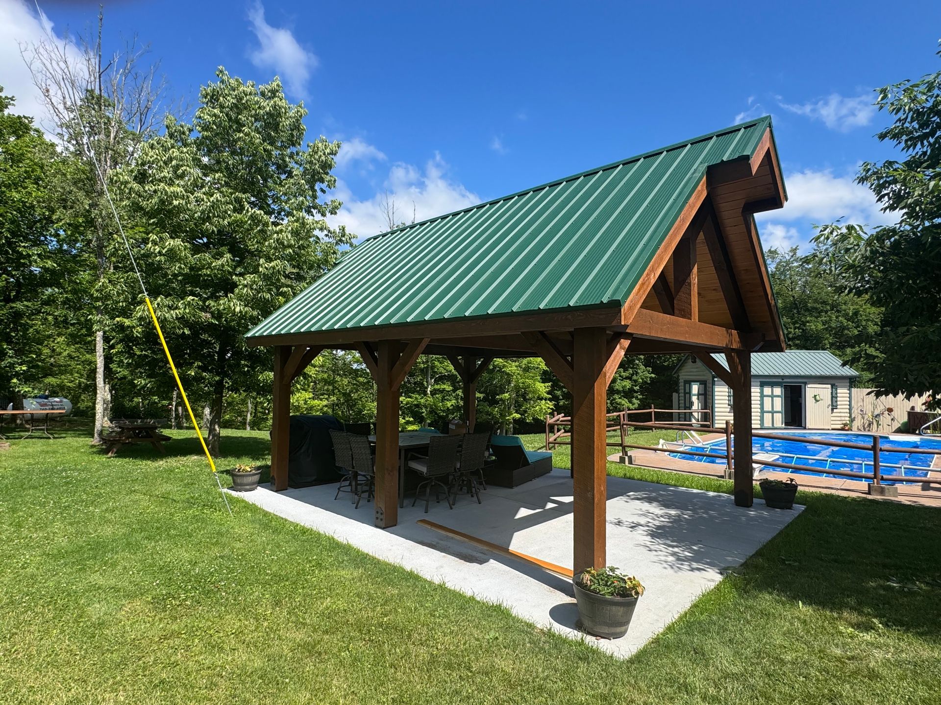 Wooden pavilion with green metal roof on a grassy lawn beside a swimming pool