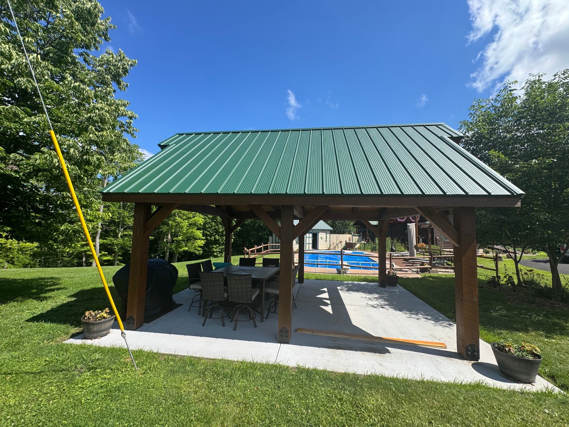 Wooden picnic pavilion with green metal roof in a sunny park, with tables underneath and trees around it