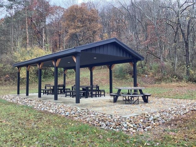 Open-sided pavilion with picnic tables on a gravel pad beside a wooded trail in fall