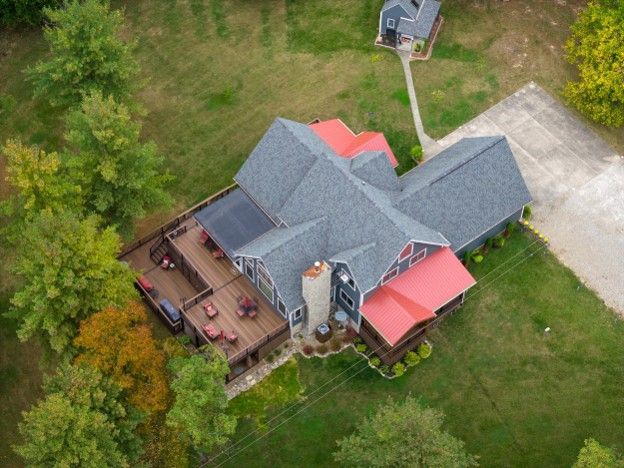 Aerial view of a large house with gray roof, red awnings, wooden deck, and surrounding grass and trees