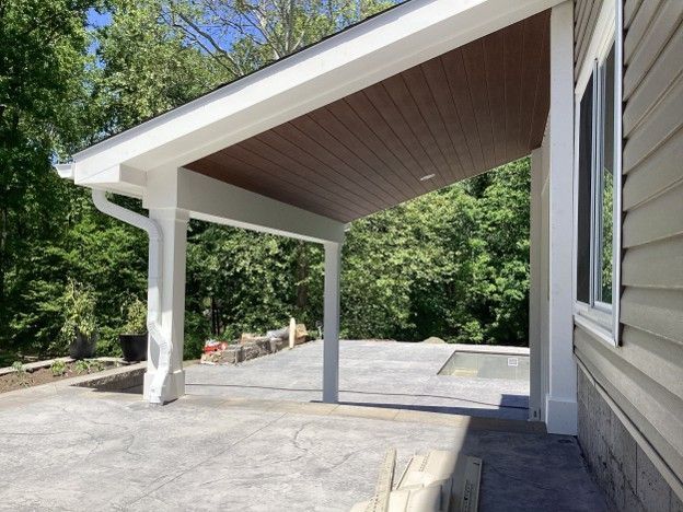 Covered patio with white support posts, sloped wood ceiling, and concrete floor beside a house and trees.