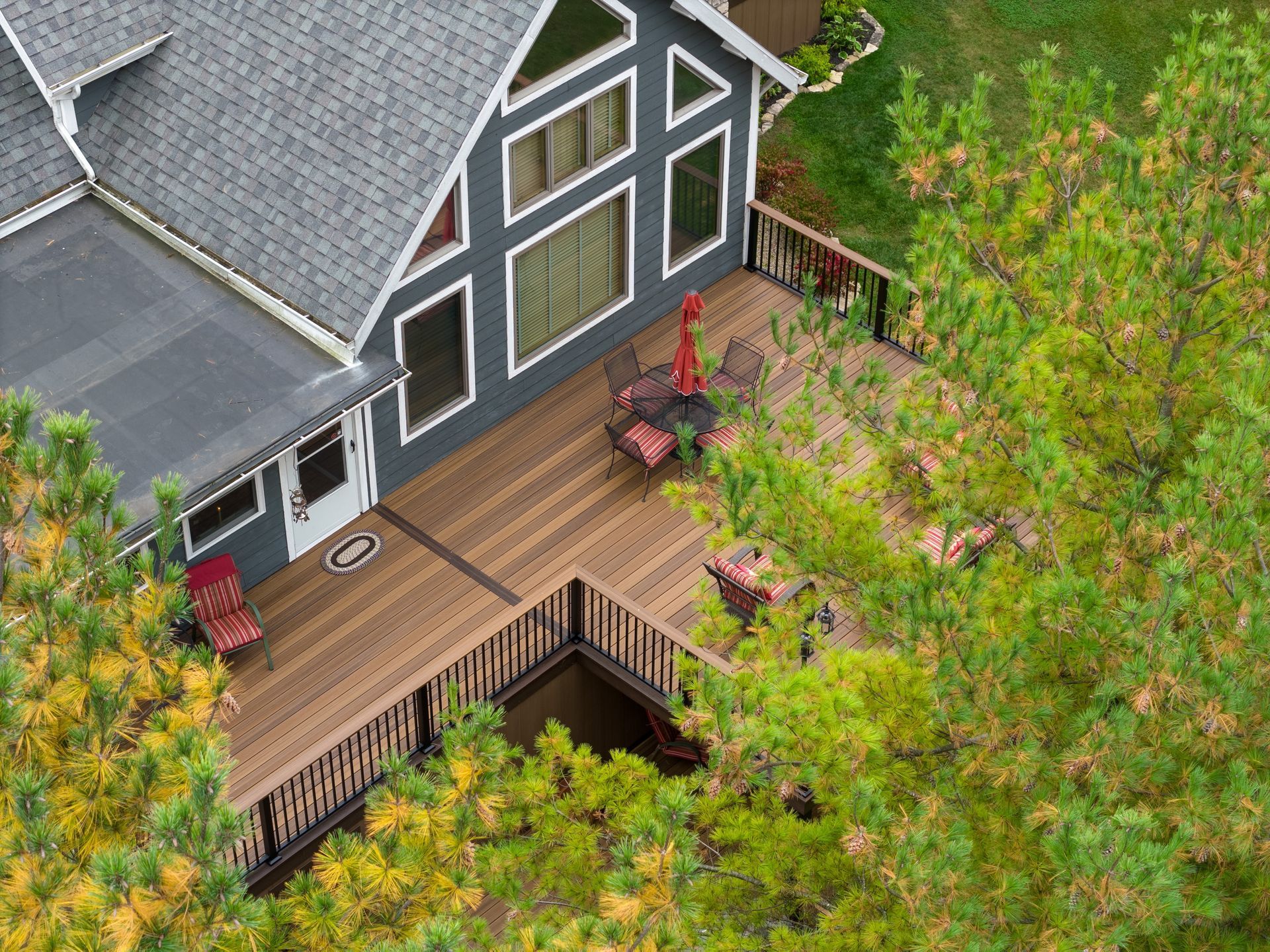 Aerial view of a gray house with a wooden deck, red chairs, and trees surrounding it