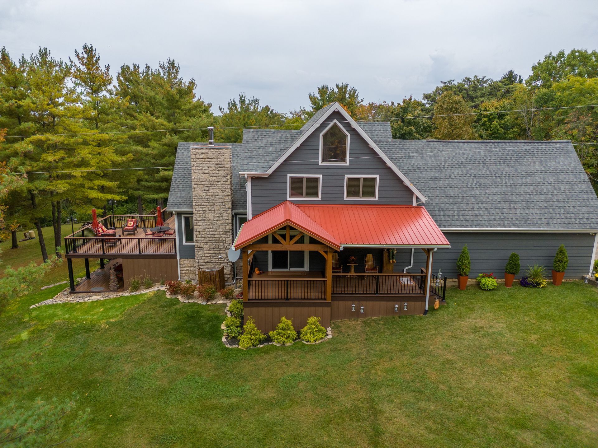 Rustic brown house with red porch roof, gray shingles, and a large grassy yard surrounded by trees