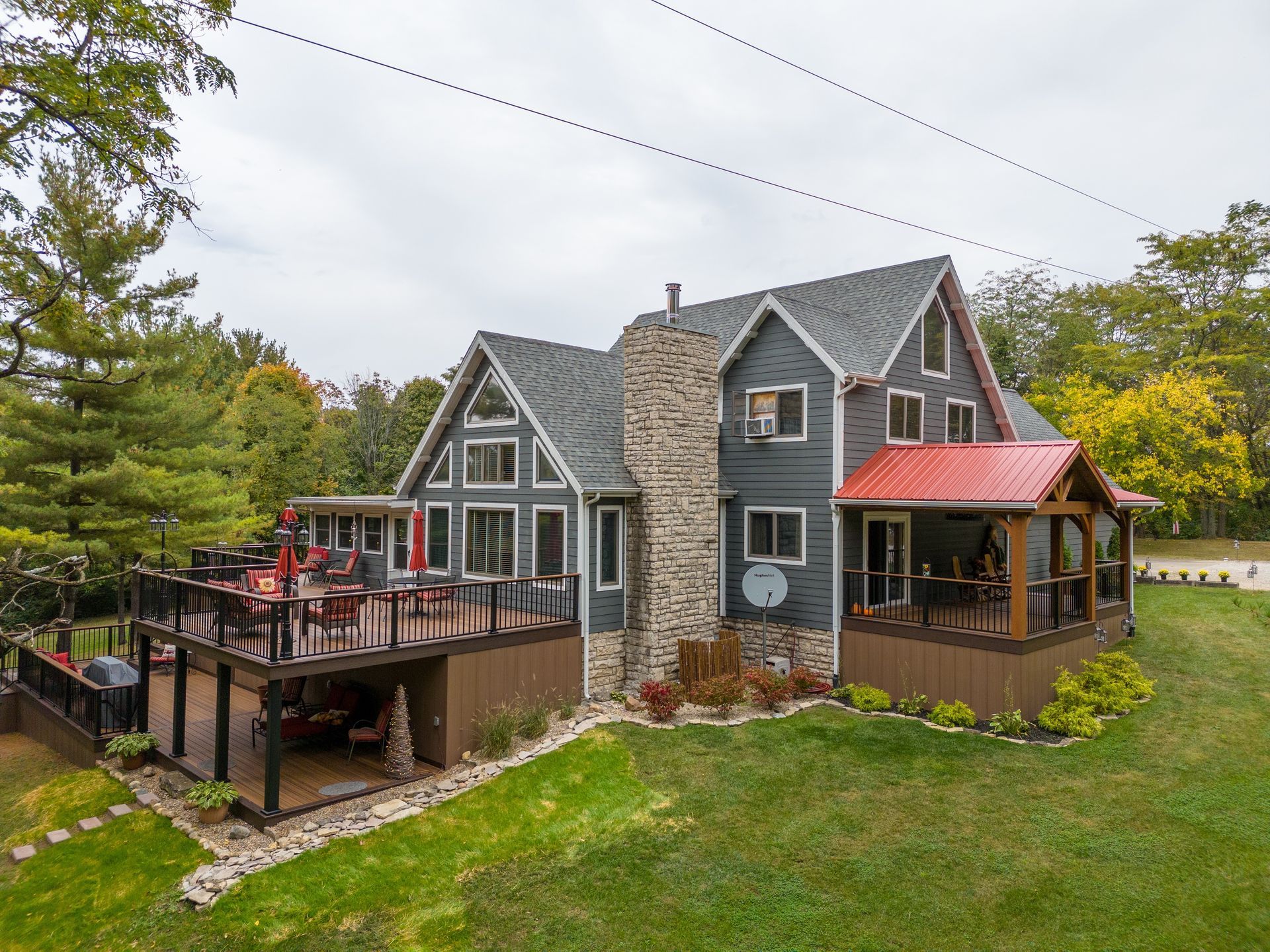 Gray stone house with red roof and deck on a grassy hillside, surrounded by trees