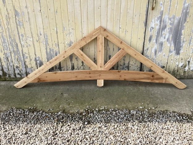 Wooden roof truss leaning against a weathered wall above gravel and concrete.