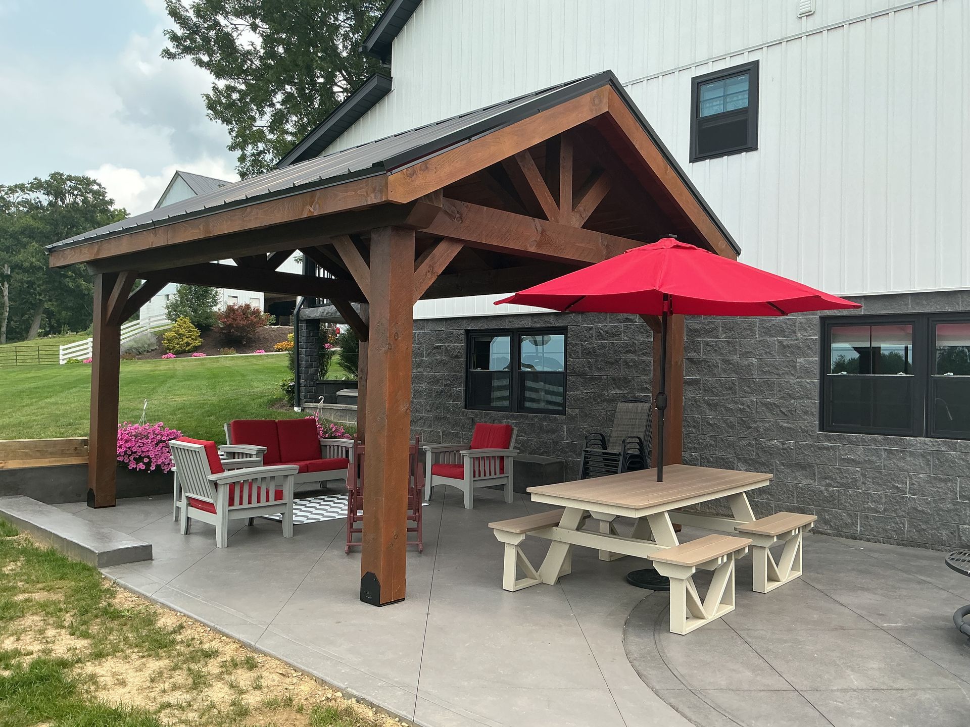 Outdoor patio with a wooden pergola, picnic table, red umbrellas, and seating beside a white building