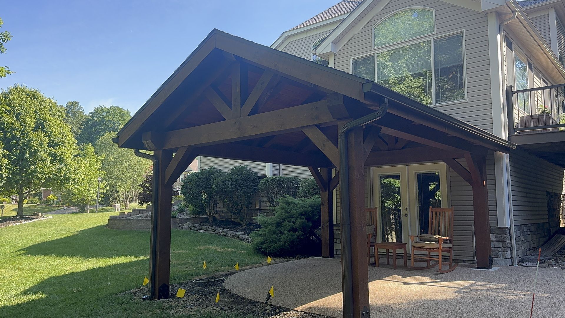 Wooden carport beside a house with stone steps, gravel ground, and a grassy yard in bright sunlight
