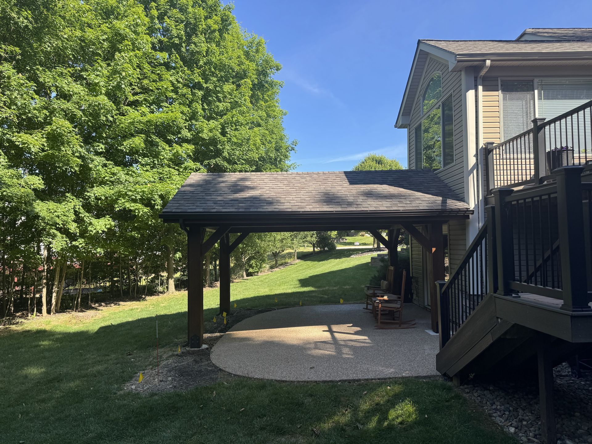 Wooden gazebo beside a house deck in a sunny backyard, with trees and a patio underneath.