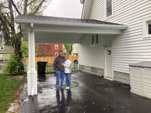 Two people standing on a wet driveway under a covered porch beside a white house.