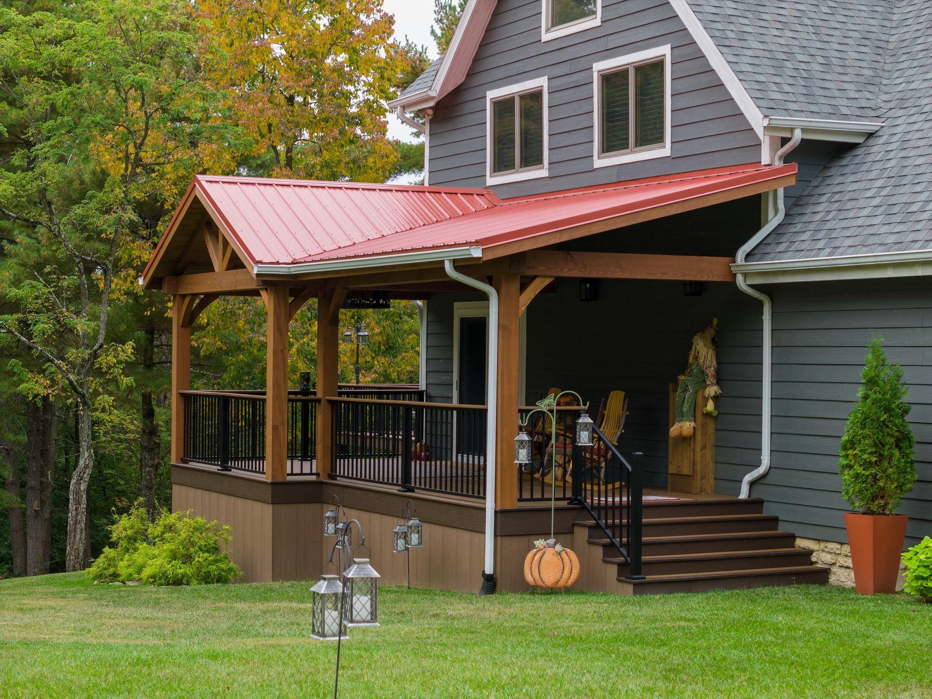 Gray house with red porch roof, steps, pumpkins, and potted topiary beside a wooded yard.
