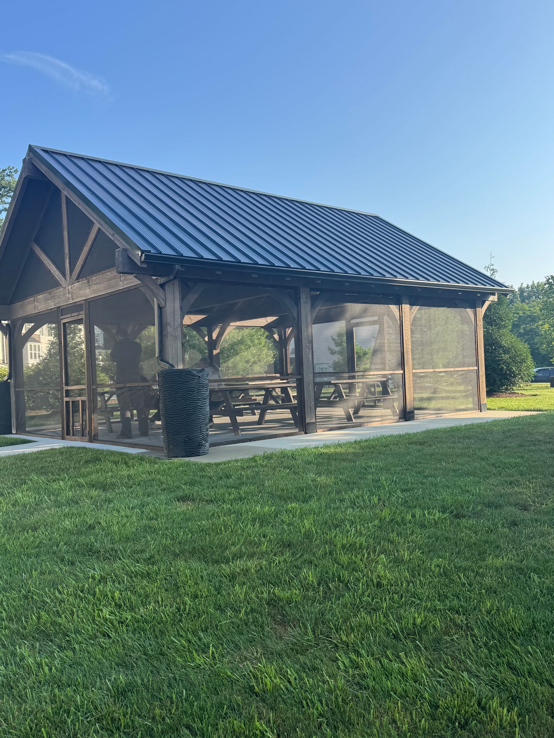 Open-sided wooden pavilion with a blue metal roof on a grassy lawn under a clear sky