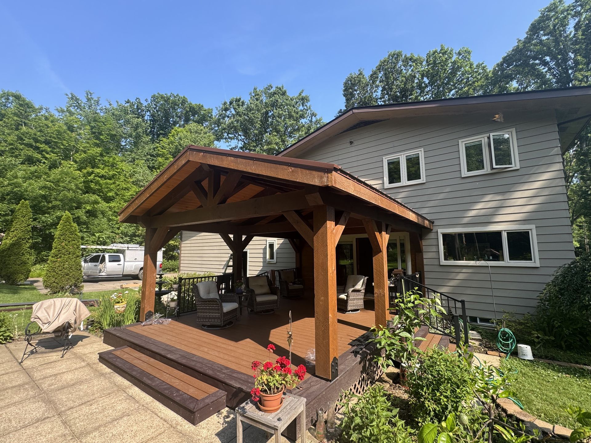 Wooden gazebo on a patio beside a gray house, with potted flowers and trees in the yard.