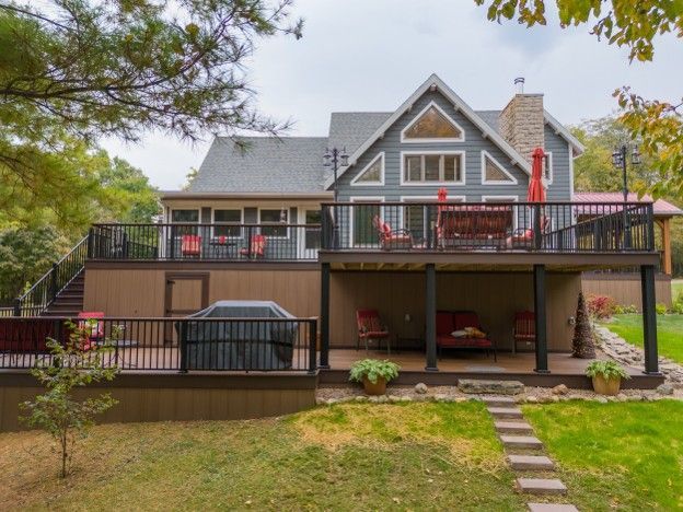 Multi-level gray house with large deck, stairs, and parked red car, viewed from a grassy yard.
