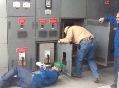 A man in a green hard hat is laying on the floor in front of an electrical panel